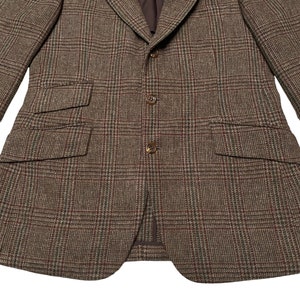 K&ouml;nnte beinhalten: Ein brauner Tweed-Blazer mit Karomuster. Der Blazer hat einen Schalkragen, einen Zwei-Knopf-Verschluss und zwei Pattentaschen.