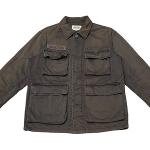 Puede incluir: Una chaqueta de trabajo marr&oacute;n oscuro con m&uacute;ltiples bolsillos y un parche que dice "AVIREX USA".