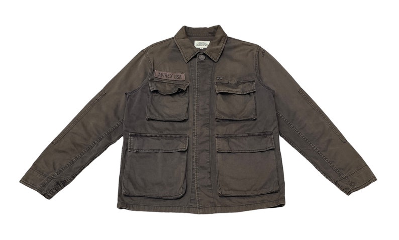 Puede incluir: Una chaqueta de trabajo marr&oacute;n oscuro con m&uacute;ltiples bolsillos y un parche que dice "AVIREX USA".