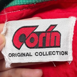 May include: A close-up of a white label with the brand name "Corin" in red, outlined in black. The text "ORIGINAL COLLECTION" is printed below the logo. The label is on a red fabric.