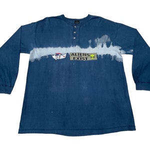 May include: A blue long-sleeve shirt with a white tie-dye pattern and the text "ALIENS EXIST" in a black rectangle with a green alien face.