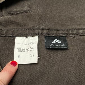 Puede incluir: Una etiqueta de prenda de algod&oacute;n marr&oacute;n con el texto "STYLE NO. 614506G" y una etiqueta de cuidado con s&iacute;mbolos para lavar, planchar y secar. La etiqueta tambi&eacute;n incluye el texto "100%" y una etiqueta negra con el texto "AVIREX&reg;".