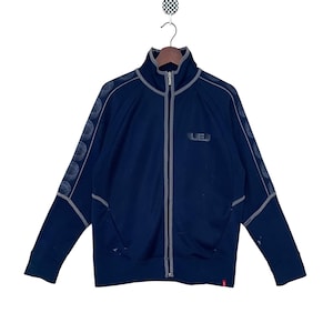 Puede incluir: Una chaqueta azul marino con cremallera que tiene una cremallera gris y ribetes grises. La chaqueta tiene un pequeño logotipo en la parte delantera que dice "LEJ".