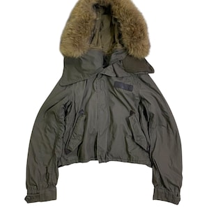 Könnte beinhalten: Olivgrüne Jacke mit brauner, fellgefütterter Kapuze. Die Jacke hat ein kurzes Design, zwei Vordertaschen und einen kleinen Aufnäher auf der Brust. Die Ärmel haben geknöpfte Manschetten. Ein stilvolles Oberbekleidungsstück.