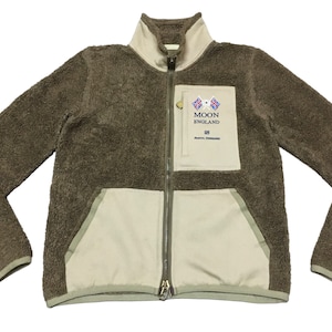 Op de afbeelding: Een bruin fleece jack met rits en een crèmekleurig paneel en een patch met de tekst "MOON ENGLAND" op de rechterborst.