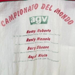 May include: White fabric with red text reading "CAMPIONATO DEL MONDO" and "agv" in a green box. Below are names: Kenny Roberts, Randy Mamola, Barry Sheene, and Angel Nieto, with championship details.