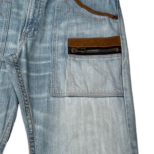 May include: Light blue denim jeans with a brown suede patch and a zippered pocket on the right side. The jeans have a red and black patterned waistband.