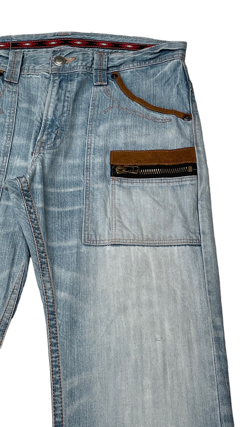 May include: Light blue denim jeans with a brown suede patch and a zippered pocket on the right side. The jeans have a red and black patterned waistband.