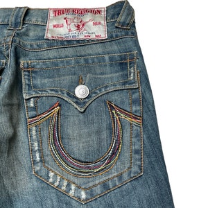 May include: A pair of blue denim jeans with a back pocket featuring a colorful embroidered horseshoe logo and a red patch with the text "True Religion Brand Jeans" and "World Tour Section Joey Bigt Row Seat".