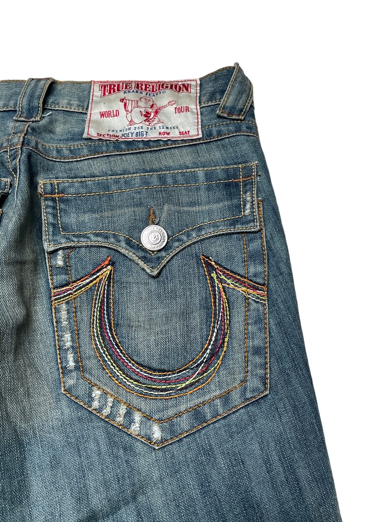 May include: A pair of blue denim jeans with a back pocket featuring a colorful embroidered horseshoe logo and a red patch with the text "True Religion Brand Jeans" and "World Tour Section Joey Bigt Row Seat".