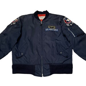 May include: A dark blue bomber jacket with a zip fastening and ribbed knit cuffs and waistband. The jacket features patches on the left and right sleeves, including a "U.S. AIR FORCE" patch on the left chest.