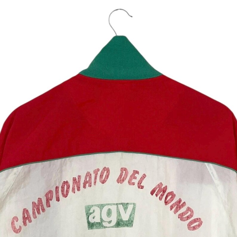 May include: A vintage red, white, and green jacket with the text "CAMPIONATO DEL MONDO" and the "agv" logo. The jacket has a green collar and is hanging on a metal hanger against a white background.