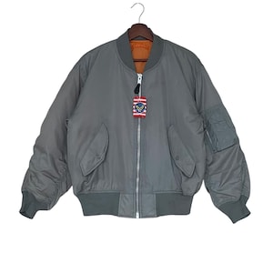Puede incluir: Una chaqueta bomber gris con cierre de cremallera y un pequeño parche en el pecho izquierdo. La chaqueta tiene un cuello y puños de canalé.