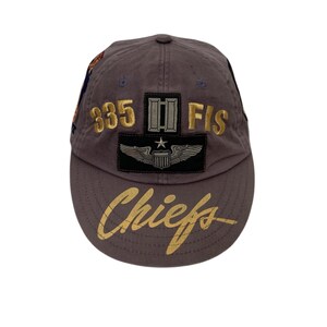 May include: A brown baseball cap with gold embroidered text and patches. The cap features the text "835 FIS" and "Chiefs" in gold, along with a patch of wings and a star. The cap is a classic baseball style.