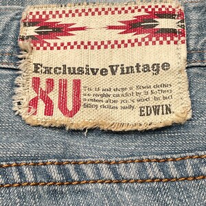 May include: A close-up of a blue denim jean with a brown stitched seam. A white fabric patch with red and black geometric designs and the text "Exclusive Vintage XU" is attached to the jean.