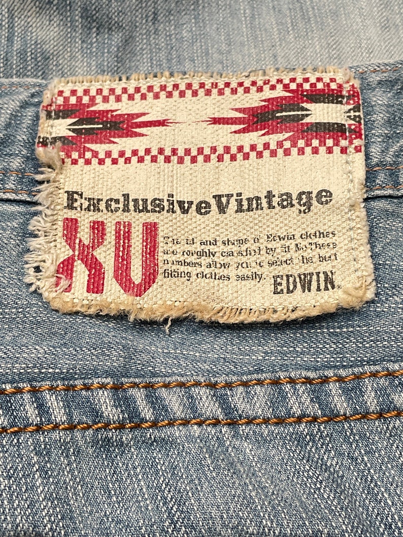 May include: A close-up of a blue denim jean with a brown stitched seam. A white fabric patch with red and black geometric designs and the text "Exclusive Vintage XU" is attached to the jean.