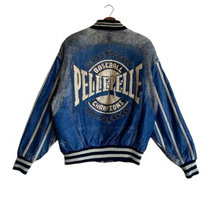 Op de afbeelding: Blauwe en grijze leren baseballjack met de tekst "BASEBALL PELLE PELLE CHAMPIONS" op de rug. De jas heeft witte strepen op de mouwen en een zwarte kraag en manchetten.