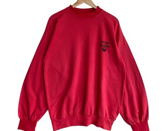 May include: A red Sergio Tacchini sweatshirt with long sleeves and a crew neck. The brand's logo is embroidered in black on the upper left chest. The sweatshirt is hanging on a wooden hanger.