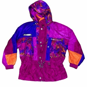 Peut inclure: Une veste de ski multicolore avec une capuche. La veste est faite d'un matériau brillant de type nylon et présente un design en patchwork avec des panneaux orange, violet et bleu. La veste a une fermeture éclair et une taille à cordon.