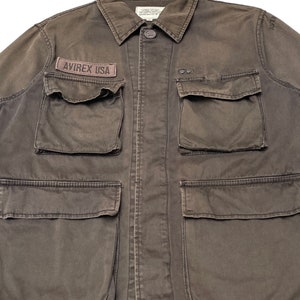 Puede incluir: Una chaqueta de trabajo marr&oacute;n oscuro con m&uacute;ltiples bolsillos y un parche que dice "AVIREX USA".