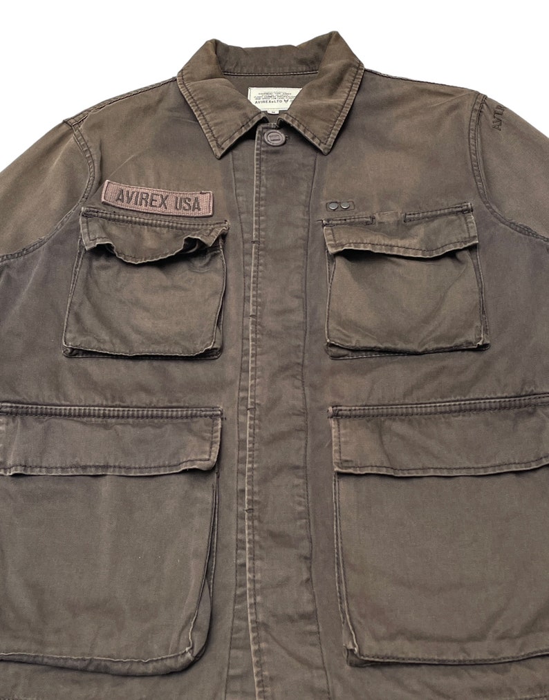 Puede incluir: Una chaqueta de trabajo marr&oacute;n oscuro con m&uacute;ltiples bolsillos y un parche que dice "AVIREX USA".