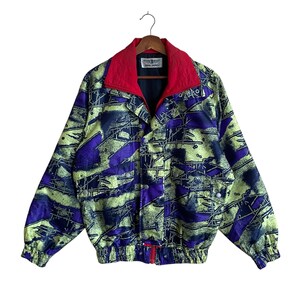 May include: A vintage jacket with a vibrant abstract pattern in shades of purple, yellow-green, and navy blue. The jacket features a red collar and a full-length zipper. The jacket has a button closure and two front pockets. The jacket is hanging on a wooden hanger.