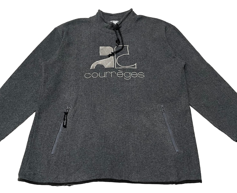 May include: A gray sweatshirt with a white logo that says "Courr&egrave;ges" and a graphic of the letter "C" with a car inside. The sweatshirt has a zippered pocket on the left side.
