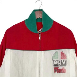 May include: A red, white, and green zip-up jacket with a green collar. The jacket features a logo on the lower right side with the letters "agv" and a design resembling a flag. The jacket is hanging on a wooden hanger.