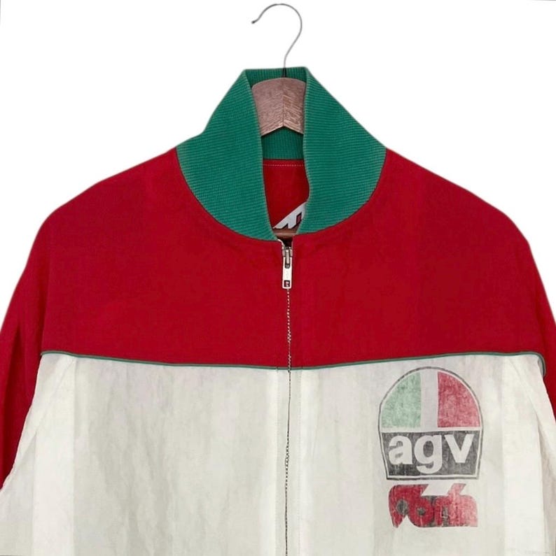 May include: A red, white, and green zip-up jacket with a green collar. The jacket features a logo on the lower right side with the letters "agv" and a design resembling a flag. The jacket is hanging on a wooden hanger.