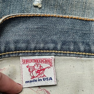 May include: A white label with the text "True Religion" in red and "made in USA" in blue. The label is attached to a pair of blue denim jeans.