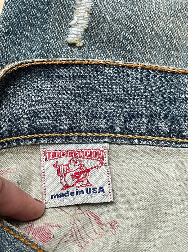 May include: A white label with the text "True Religion" in red and "made in USA" in blue. The label is attached to a pair of blue denim jeans.