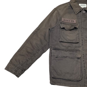 Puede incluir: Una chaqueta de trabajo marr&oacute;n oscuro con un parche que dice "AVIREX USA" en el pecho izquierdo. La chaqueta tiene varios bolsillos y cierre de botones.