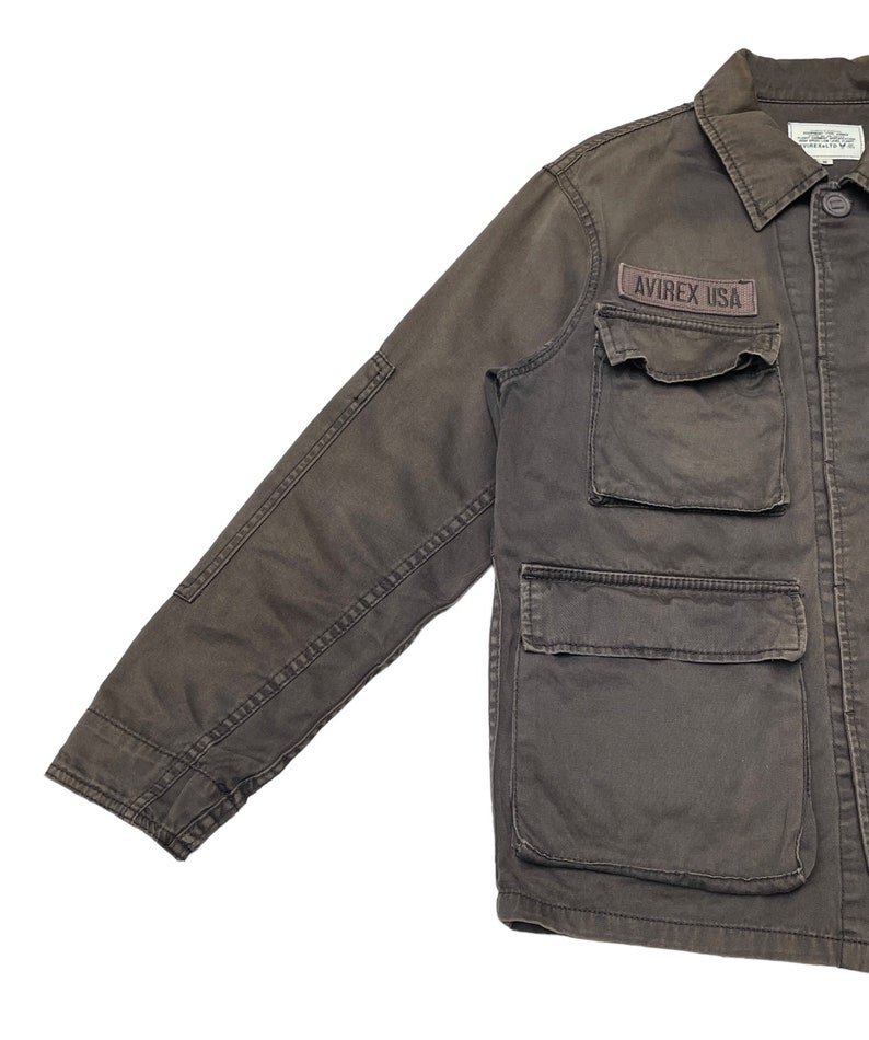 Puede incluir: Una chaqueta de trabajo marr&oacute;n oscuro con un parche que dice "AVIREX USA" en el pecho izquierdo. La chaqueta tiene varios bolsillos y cierre de botones.