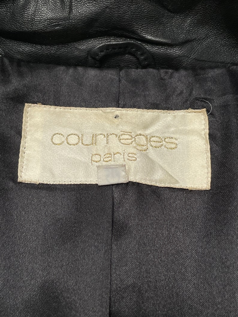 May include: A white label with the text "Courr&egrave;ges Paris" in gold lettering. The label is attached to a black leather jacket.