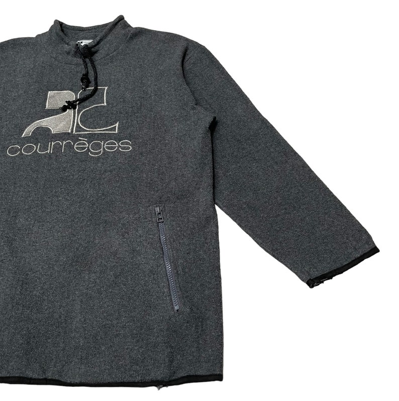 May include: Gray fleece pullover sweatshirt with a black zippered pocket on the left side. The sweatshirt has a high neck with a drawstring and features a white embroidered logo that says "2C courr&egrave;ges".