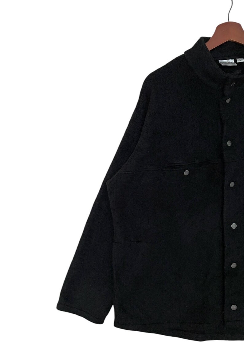 May include: A black fleece jacket with a button-up front and a single chest pocket. The jacket has a stand-up collar and long sleeves.