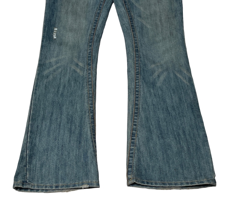 May include: A pair of blue denim jeans with a flared leg style. The jeans have a distressed look with a small hole on the left leg.