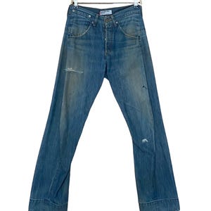 Puede incluir: Un par de pantalones vaqueros azules de mezclilla con detalles desgastados, colgados de una percha de madera. Los pantalones tienen una pierna recta y un dobladillo con vuelta.