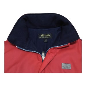 May include: Red jacket with a blue collar and a zipper closure. The jacket has a label that says "Yamaha for Sports Enthusiasts" and a small patch on the lower right side.