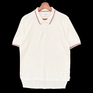 May include: White short-sleeved polo shirt with a red and blue striped collar and cuffs. The shirt has a button-up closure and a small label on the bottom.
