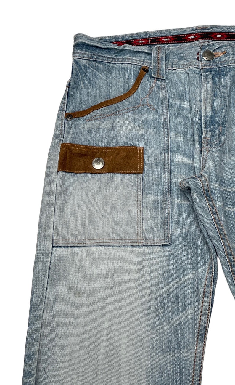 May include: Light blue denim jeans with a brown suede patch pocket and a red and black patterned waistband.