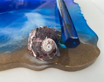 May include: A decorative resin tray with a beach theme. The tray features a seashell on a sandy base, with blue and white resin simulating ocean waves. A blue, geometric accent piece adds a modern touch.