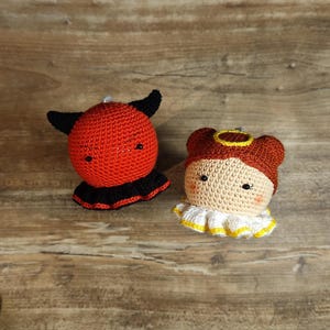 Puede incluir: Dos muñecos de ganchillo: un diablo rojo con cuernos negros y un cuello negro, y un ángel marrón claro con un halo y un cuello blanco y amarillo. Los muñecos están hechos a mano y están sobre una superficie de madera.