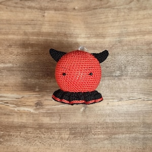 Puede incluir: Adorno de diablo rojo hecho a mano con ganchillo, con cuernos negros y un cuello con volantes negro. El pequeño adorno redondo tiene dos pequeños ojos negros y está suspendido por un hilo transparente. Fondo de superficie de madera marrón claro.