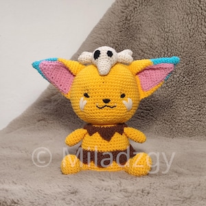 May include: A crocheted yellow and brown plush toy with a white skull on its head. The toy has pink ears and blue accents. The toy is sitting on a light brown fluffy surface.