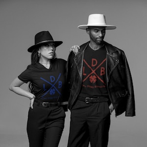 May include: Two people wearing black t-shirts with the text "LDB" and a four-leaf clover design. One shirt has blue text, the other red. Both are wearing black pants, belts, and hats. One person wears a black leather jacket.