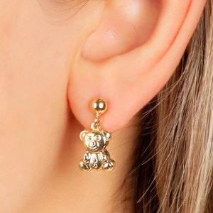 May include: Gold-colored bear-shaped dangle earrings. Each earring features a small, detailed teddy bear pendant suspended from a round stud. The earrings are shown on a person's ear, with the focus on the jewelry.