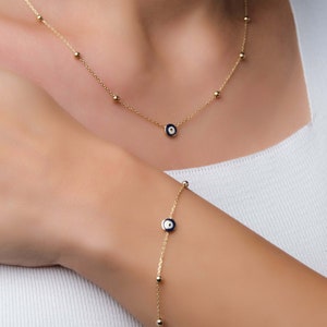 以下が含まれることがあります： 青と白の邪悪な目をモチーフにした金のネックレスとブレスレット。ネックレスとブレスレットはどちらも、小さな金のビーズが付いた繊細なチェーンで作られています。