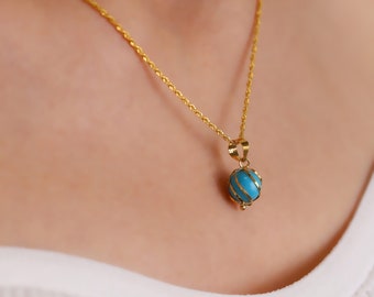 May include: A gold necklace featuring a turquoise pendant. The pendant is a sphere encased in a gold cage design. The necklace is worn against a white top.