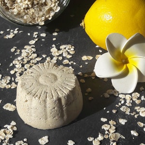Puede incluir: Una pastilla de jabón redonda, texturizada, de color marrón claro con un diseño floral sobre una superficie negra con copos de avena esparcidos.